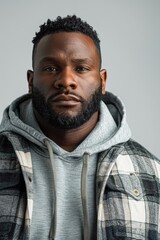 A young man with a beard and a serious expression looks directly at the camera. He wears a gray hoodie under a plaid flannel shirt