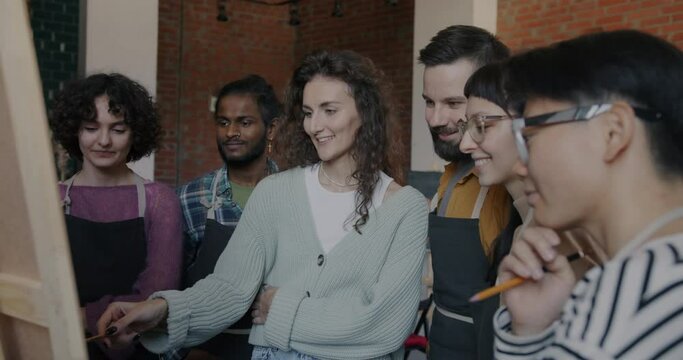 Female artist teaching diverse group of students pointing at picture and speaking in class. Professional occupation and youth culture concept.