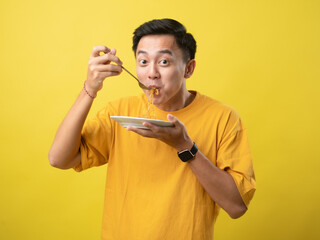 A young man in a yellow shirt is enthusiastically eating a plate of noodles with a fork against a...