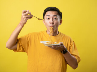 A young man in a yellow shirt is enthusiastically eating a plate of noodles with a fork against a...