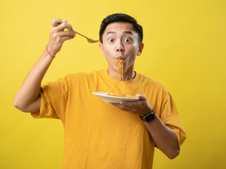 A young man in a yellow shirt is enthusiastically eating a plate of noodles with a fork against a...