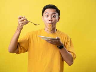 A young man in a yellow shirt is enthusiastically eating a plate of noodles with a fork against a...
