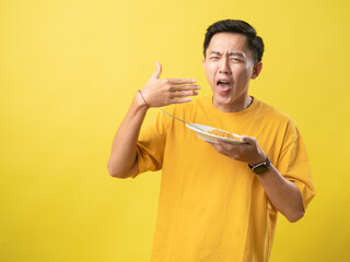 A young man in a yellow shirt reacts to hot or spicy noodles on a plate, using a fork, against a bright yellow background.