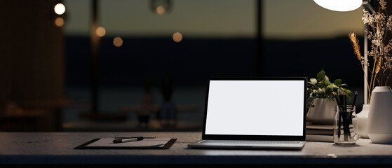 A modern dark office at night featuring a white-screen laptop computer mockup on a table.