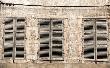 three heavily weathered windows with closed blinds stand in line on an old, weather beaten wall