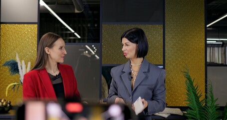 Interior design masterclass by professional bloggers. Two women in elegant outfits review fabrics and create mood boards, filming online tutorial with smartphone. Engaging, trendy, educational content