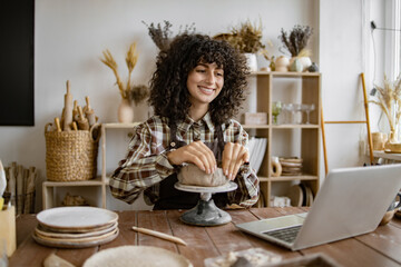 Young woman shaping clay on pottery wheel while watching online tutorial on laptop in ceramic studio