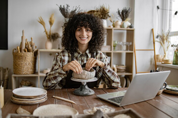 Young female artist creating pottery at wooden table in cozy studio with laptop. She is smiling and...
