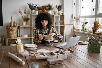 Young woman crafting pottery in cozy home studio with laptop on wooden table. Creative and artistic...