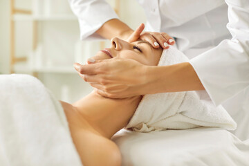 Female masseur making face relaxing massage to a young girl lying with closed eyes in beauty salon or cosmetology cabinet. Professional cosmetologist making massage for woman in spa.