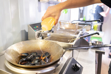 A hand pours liquid from a bottle into a frying pan with mussels on a stove, suggesting cooking or...