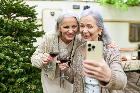 A middle-aged lesbian couple enjoys a glass of wine and takes a selfie in the forest while camping.