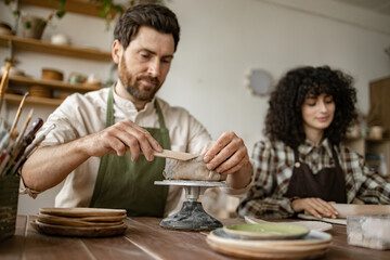 Bearded mature man in apron sculpts vase while a woman rolls clay with a rolling pin in pottery studio.