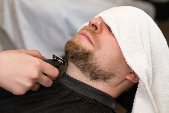 The clients head is covered with a towel during a beard trim. Shortening the length of the beard from the sides by the master for the client.