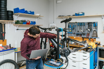 The bike mechanic is manipulating and repairing an electric mountain bike while holding a laptop with the other hand, likely displaying a tutorial or checklist for the bike.