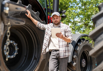 Man farmer stands next to an agriculture tractor and using his digital tablet. Agricultural...
