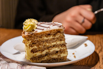 Slice of tasty homemade delicious sweet Esterhazy cake with nuts and cream. Piece of Esterhazy Torte served on the white plate with napkin. Young woman´s hand on the blurred background Selective focus