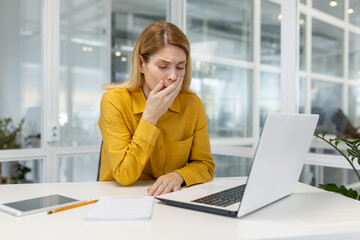 Tired businesswoman yawning while working on a laptop in a modern office, showing fatigue and exhaustion during a busy workday.