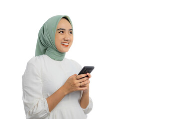 Happy young Asian woman in green hijab and white blouse holding mobile phone and looking away with friendly smile isolated on white background