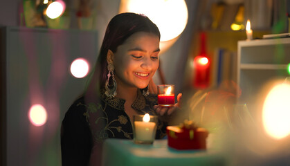 Indian beautiful happy teen age girl smiling sitting indoor decor home hold red glass burn candle...