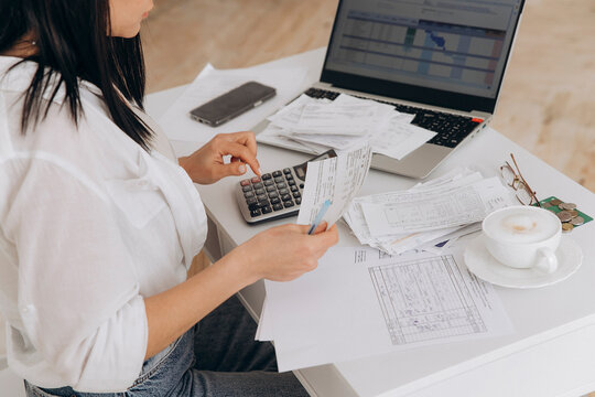 Unrecognizable woman managing financial documents on white office desk with laptop, using a calculator for bill payments and budget planning. Independent accounting, remote financial management
