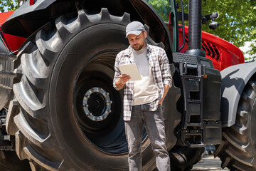 Man farmer stands next to an agriculture tractor and using his digital tablet. Agricultural mechanic searching information on the internet.