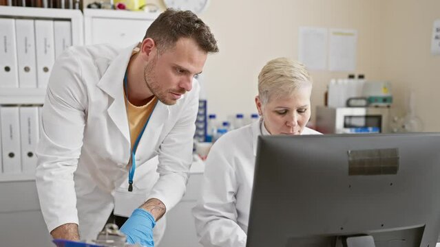A man and a woman in lab coats analyze data on a computer in a laboratory setting, reflecting teamwork and research.