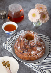 A French Klouglof in a plate with flowers, knife and spoon in background with forks, pot and cup of tea with flowers. It's a cake traditionally baked in a distinctive ring pan.