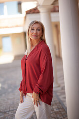 Beautiful blond middle-aged woman dressed red shirt walking in the street. Outdoor portrait of a 55 - 60 s ears old woman. Close up of fashion urban outfit