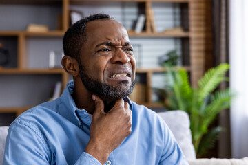 A man sitting at home, experiencing discomfort or pain in his throat, and touching his neck. The image conveys feelings of illness, discomfort, or medical condition.