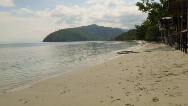 A tranquil beach on Kri Island, Raja Ampat, Indonesia. The serene waters gently lap against the pristine shore, while two-masted yacht is anchored  in a distance. Pan left shot.