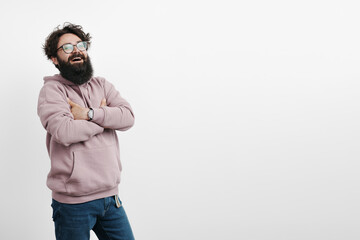 A joyful bearded man in a pink hoodie laughs with his arms crossed, standing against a white...