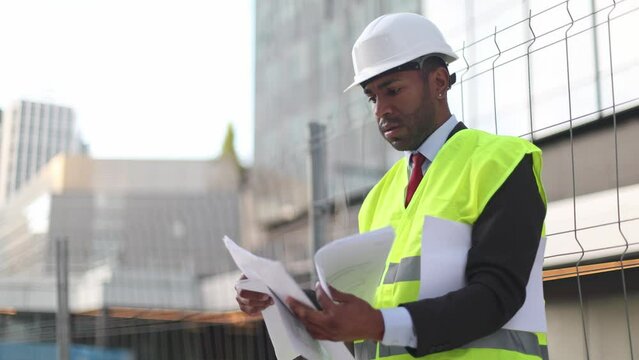 Architect in hard hat and safety vest reviewing documents at construction site.