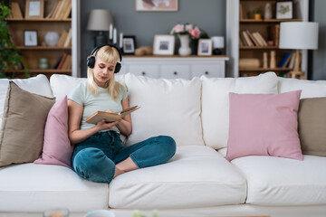 Young millennial woman sitting at home on sofa listening music on wireless headphones read book enjoy life and freedom. Educated university student girl resting and relaxing with mix tape and reading.