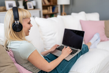 Attractive young woman sitting cross legged on sofa using her laptop and headphones to make video...