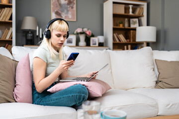 Attractive young woman sitting cross legged on sofa using her laptop and headphones to make video call chat at home. University student girl online dating on computer, listening music, working at home