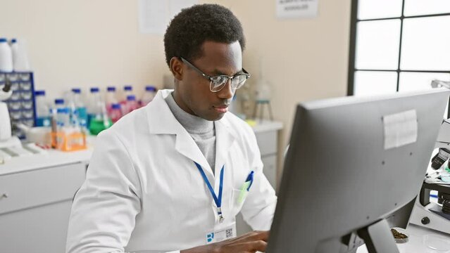 A focused african man in a white lab coat works on a computer in a laboratory setting.