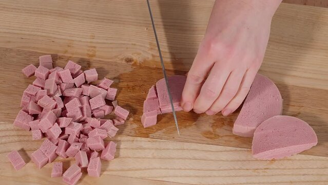 The sausage is cut into cubes on a wooden board with a sharp knife. Ham filling for breakfast. Appetizers with ham. Sandwiches, scrambled eggs. Close-up, macro, top view