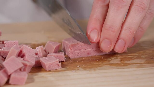 The chef cuts the ham into cubes with a sharp knife on a wooden board. Sausage for breakfast, preparations for eggs. Appetizers with ham. Close-up, macro, front view