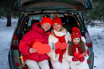 Family - young father mother and daughter sitting in the open trunk of a car holding red hearts in their hands in a snowy winter coniferous forest