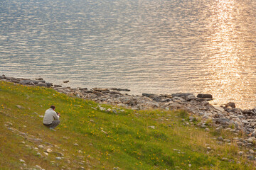 Baikal Lake in June at dawn. Tourist photographs yellow iris flowers on green meadow on beach of...