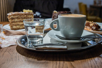 Cup of coffee latte with cream foam served on the white plate with spoon. Few plates with cakes and desserts, glass of water, sugar on a metal tray on the wooden table in coffee shop. Selective focus
