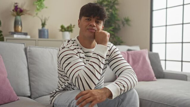 Young hispanic man in a striped shirt sits in a modern living room, casually posing in front of a sofa with pink and gray cushions, surrounded by plants and stylish decor.