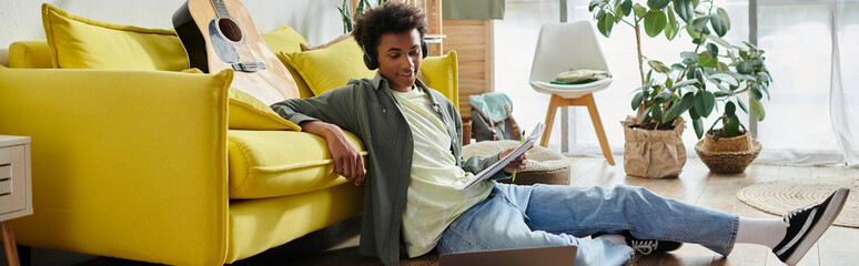 A young African American man sits on a yellow couch, focused on his laptop screen.