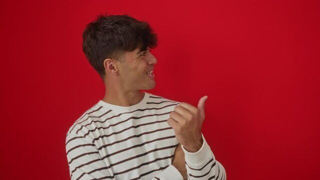 Young hispanic man wearing stripes t shirt standing smiling with happy face looking and pointing to the side with thumb up. over isolated red background
