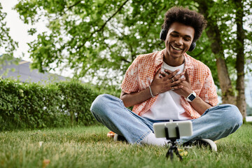 Obraz premium Young African American man sitting on grass, engrossed in cell phone.