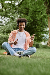 Obraz premium Young African American man sitting on grass, pointing at his phone.