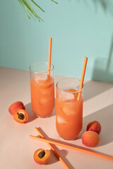 Two glasses with iced apricot cocktail beverages, fruits and peachy drinking straws on bright pink and turquoise background with aesthetic sunlight shadows, old fashioned style
