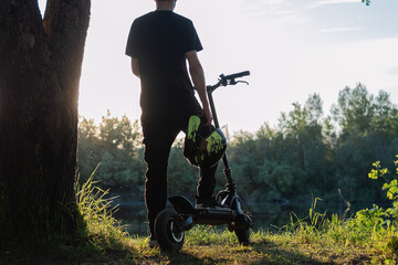 man on an electric scooter stands with his back to the sunset, holding a helmet