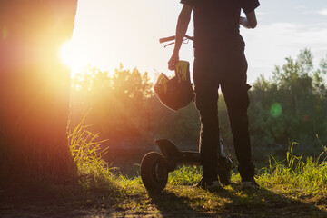 man on an electric scooter stands with his back to the sunset, holding a helmet